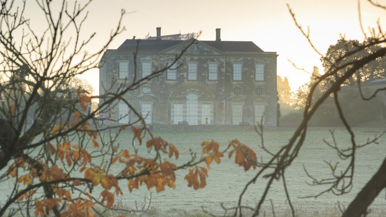 A grand house on a grassy bank illuminated by the sun rising behind it and slightly obscured by the last orange leaves of autumn on the trees in the foreground.